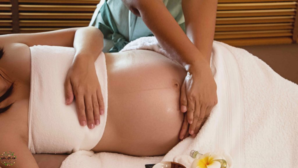 زن بارداری به پهلو بر روی تخت ماساژ دراز کشیده و ماساژور زیر شکم او را ماساژ بارداری میدهد، زن باردار بدن خود را با حوله های سفید پوشانده است