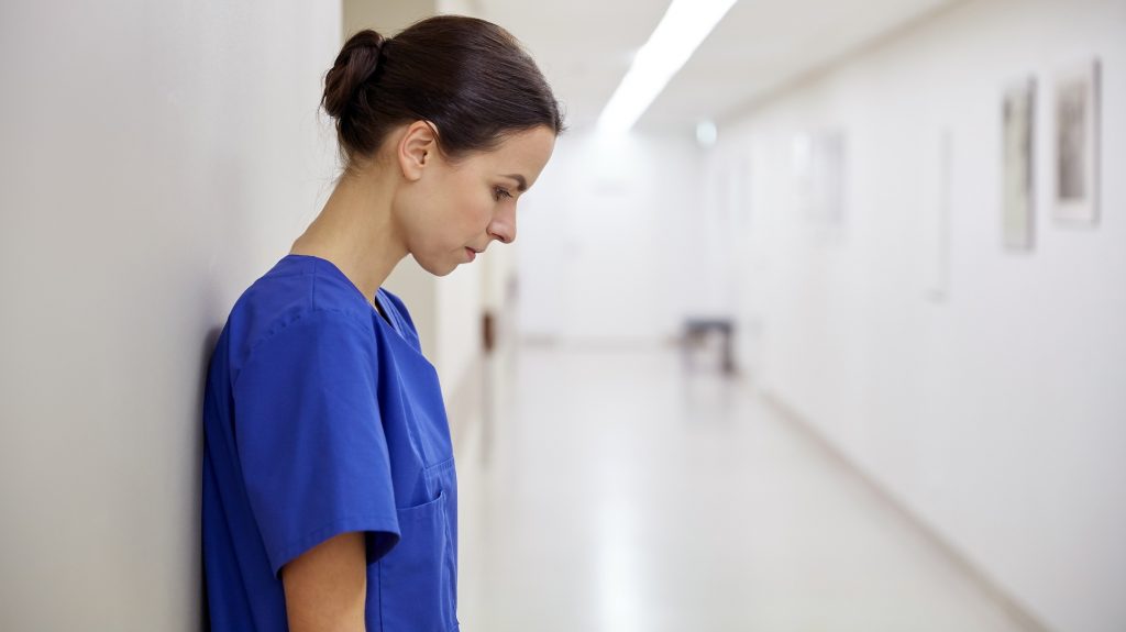 پرستار زن غمگین در بیمارستان به دیوار تکیه داده است و به فکر فرو رفته، لباس پرستاری آبی پوشیده است