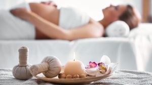 زن بارداری بر روی تخت ماساژ دراز کشیده و در حال آروماتراپی است، در کنار او شمع ، گل و حوله قرار دارد