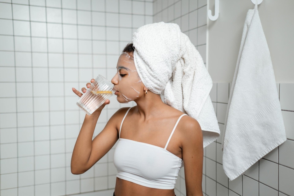 زن جوان با حوله‌ای بر سر، در حال نوشیدن آب پس از ماساژ در یک حمام روشن